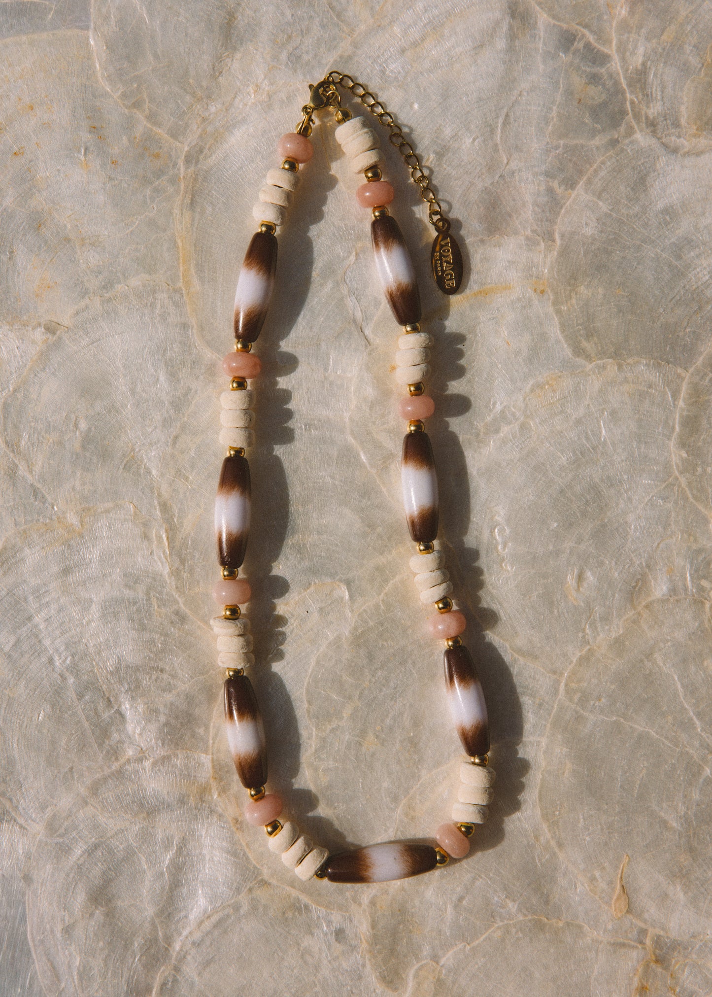 A bohemian-style beaded necklace featuring brown and white marbled beads, cream cylindrical beads, small pink beads, and gold spacer rings, laid flat on a textured beige stone surface.