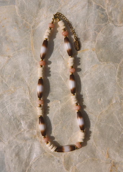 A bohemian-style beaded necklace featuring brown and white marbled beads, cream cylindrical beads, small pink beads, and gold spacer rings, laid flat on a textured beige stone surface.