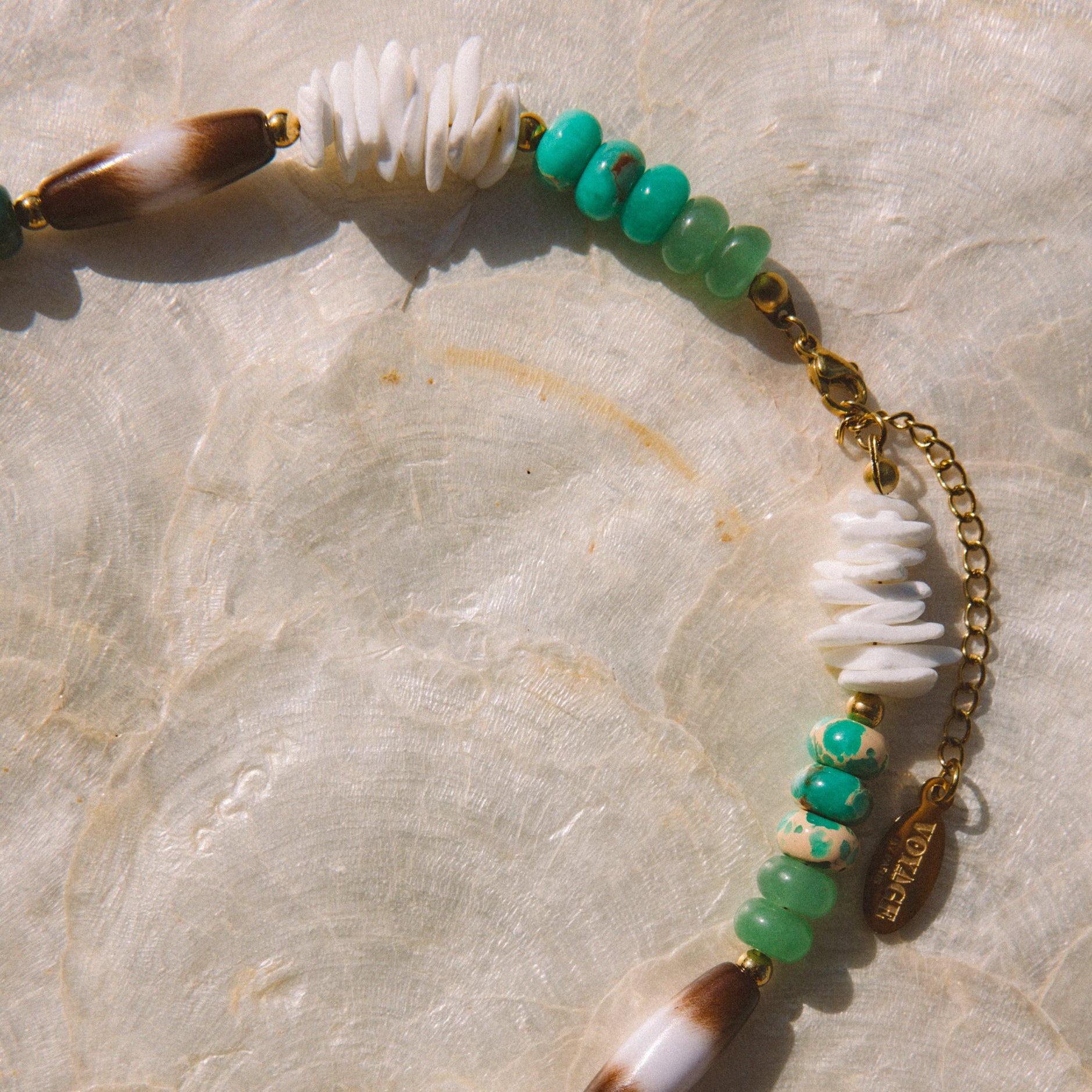 A bohemian-style beaded necklace featuring white beads, turquoise and green beads, and gold-toned metal accents, laid flat on a textured beige stone surface.