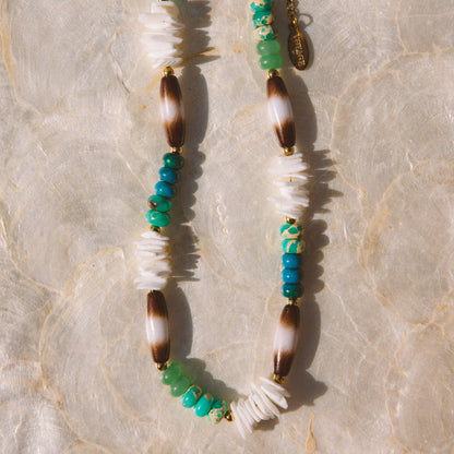 A bohemian-style beaded necklace featuring turquoise, white, brown, and gold beads, laid flat on a textured beige surface.