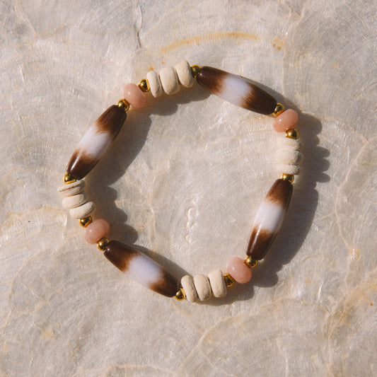 A beaded stretch bracelet featuring alternating white and brown marbled beads, small pale pink beads, and gold spacer beads, displayed on a textured beige stone surface.