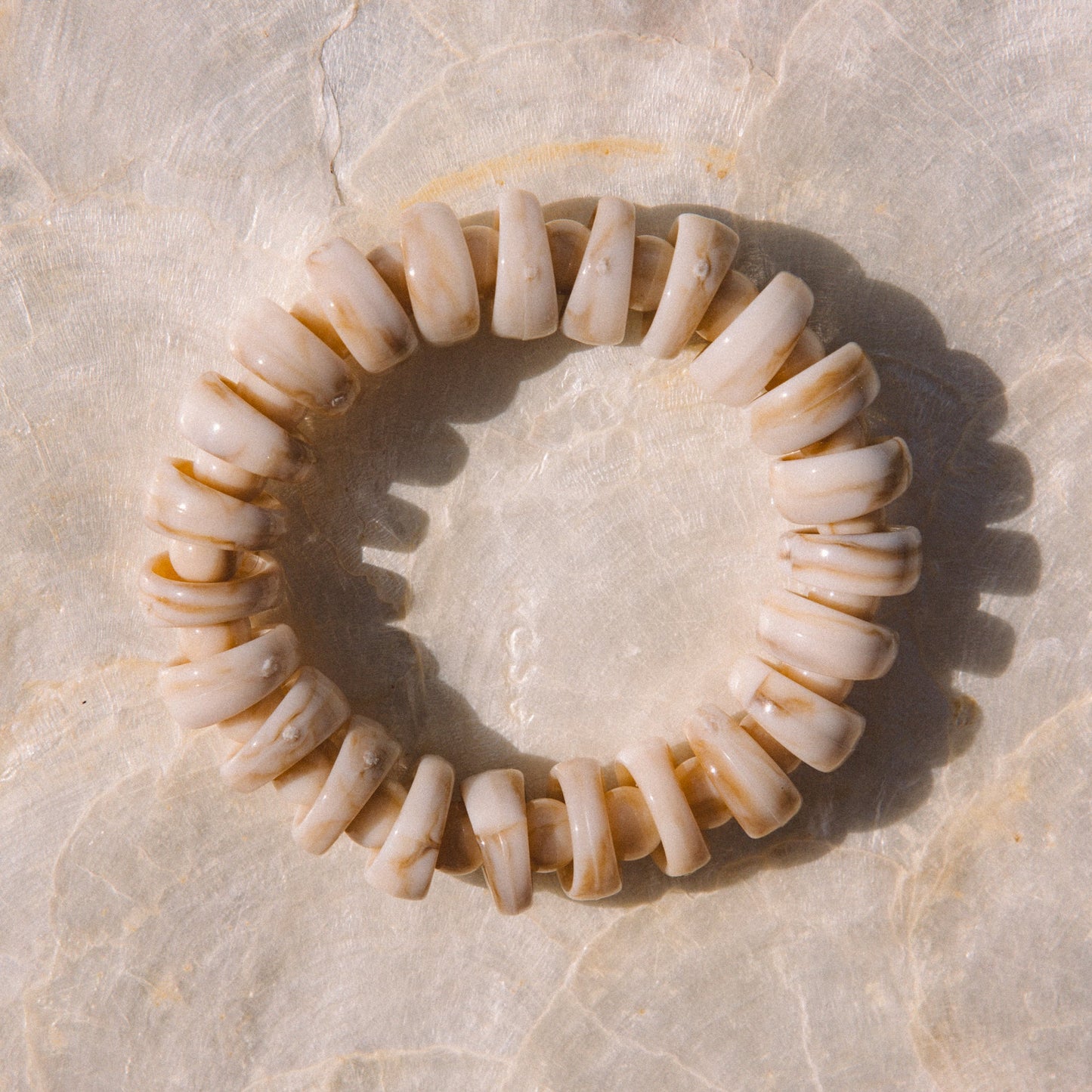 A beaded anklet made of small, rounded acrylic pieces in natural cream and light brown tones, arranged in a circle on a light beige marble surface.