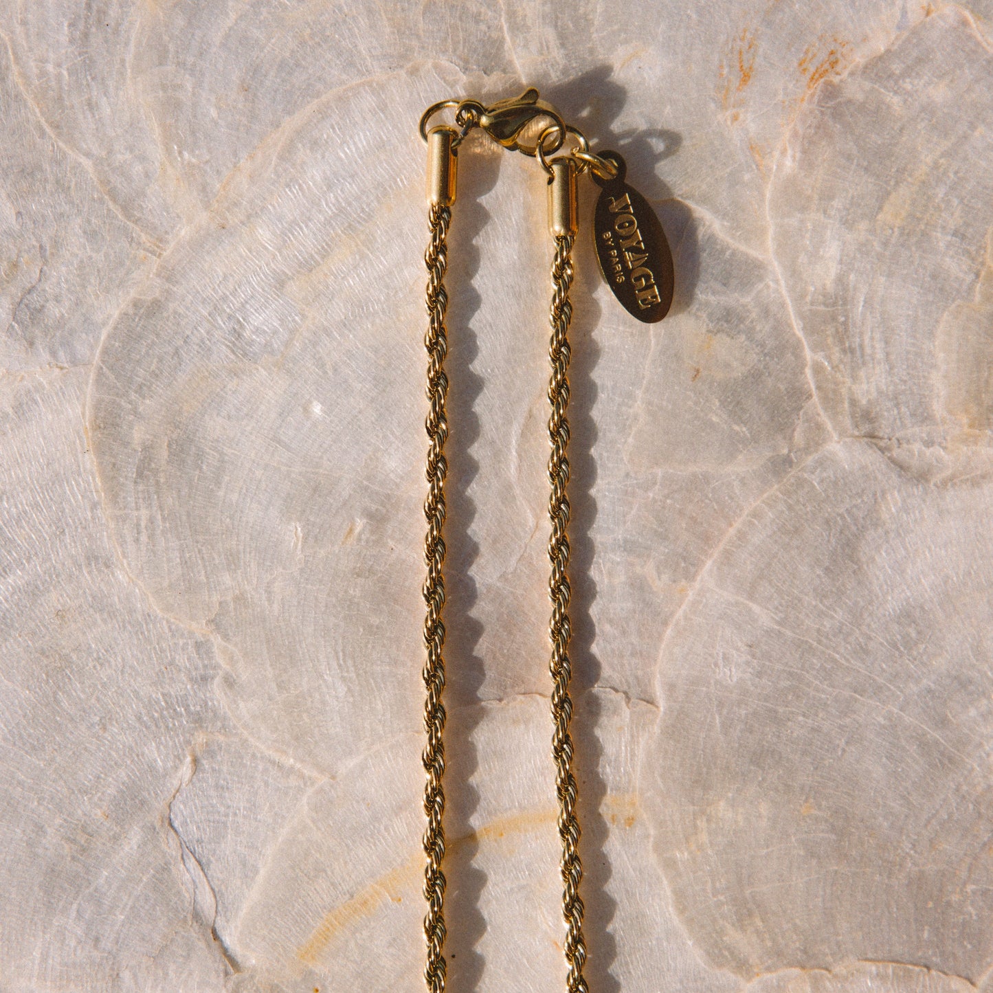 A close-up of a gold chain necklace with a lobster clasp and a small hanging tag, laid on a light beige stone surface.
