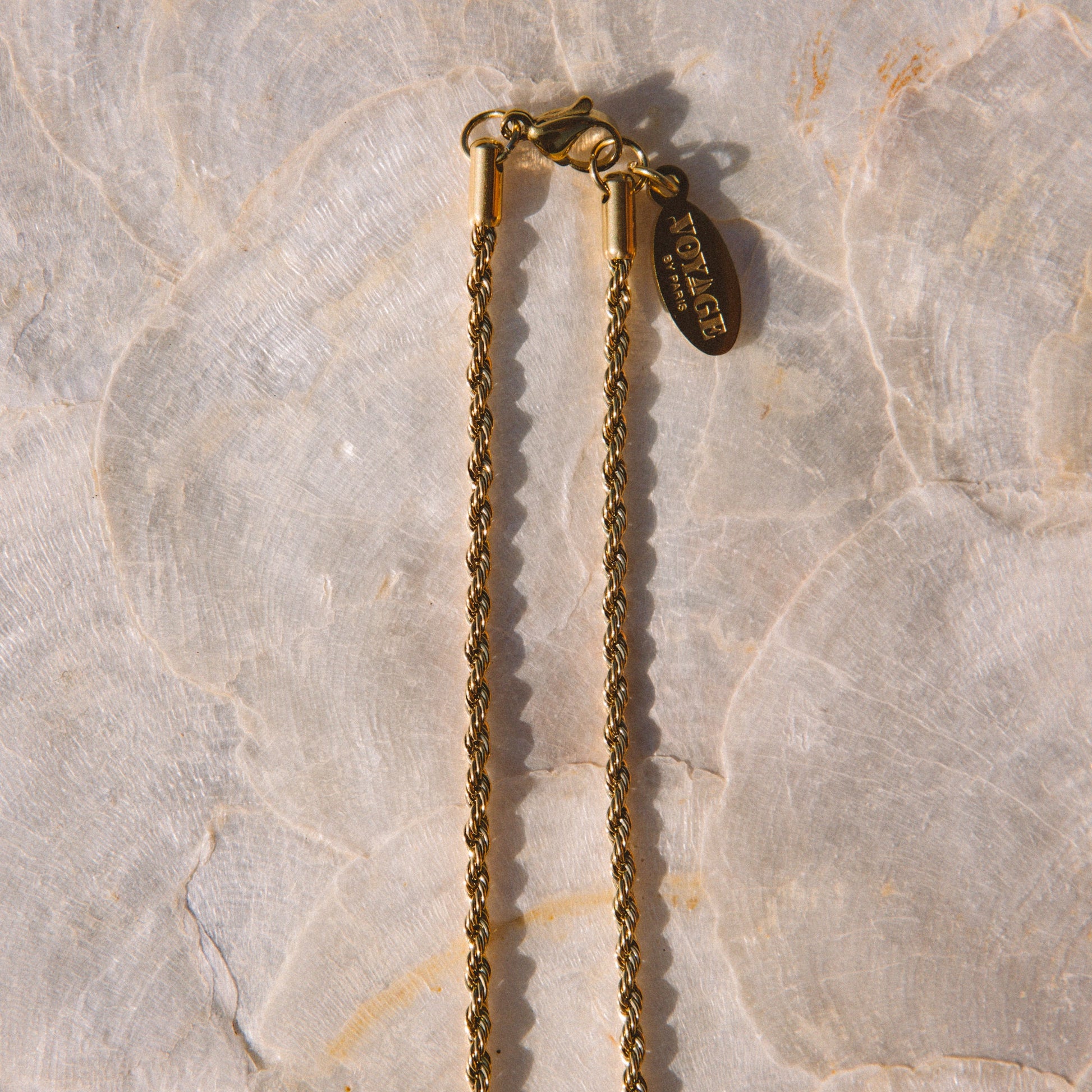 A close-up of a gold chain necklace with a lobster clasp and a small hanging tag, laid on a light beige stone surface.