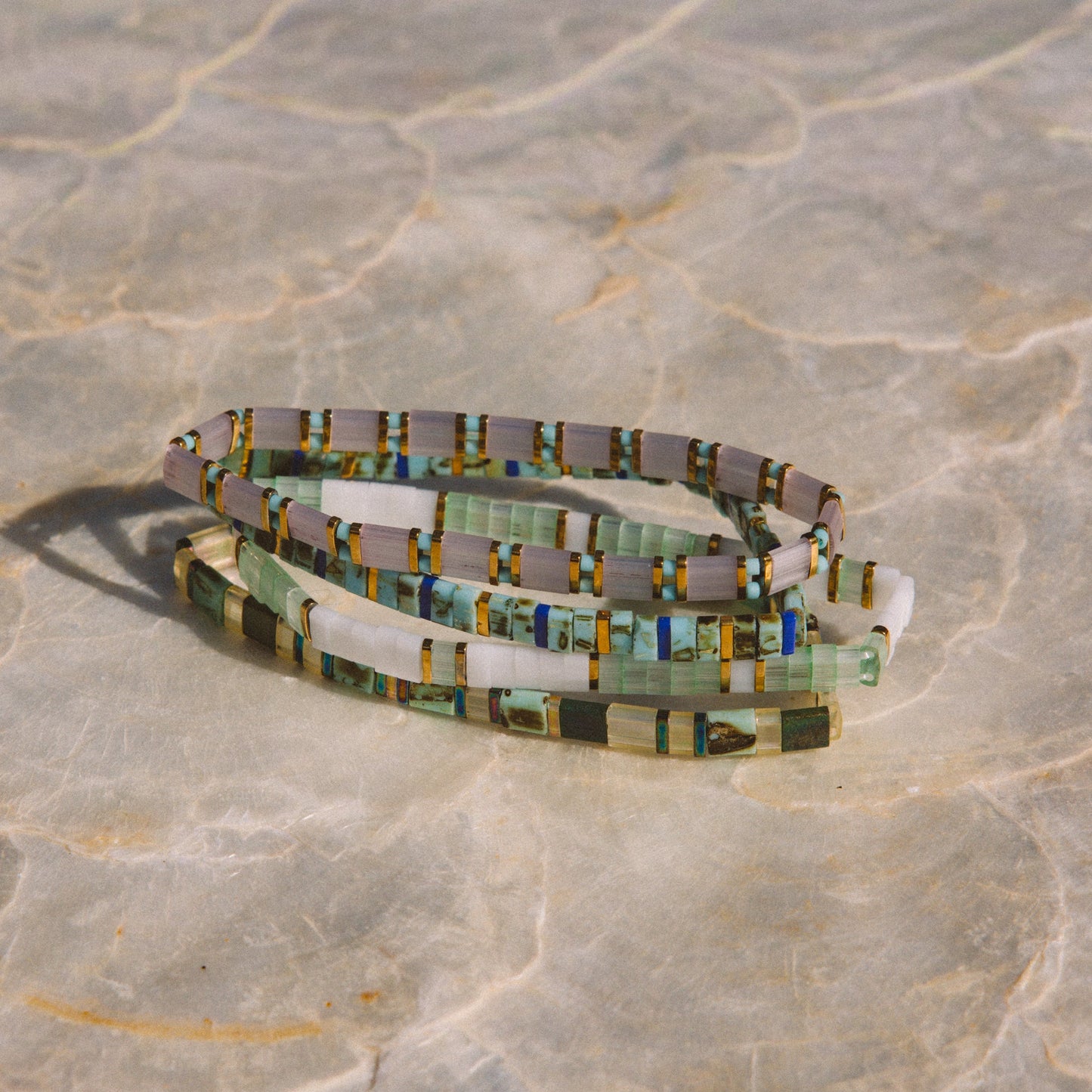 A coiled beaded bracelet or anklet made of small rectangular glass pieces in muted green, gray, white, and touches of yellow and blue, photographed on a light stone-like surface.