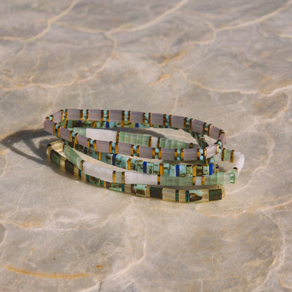 A coiled beaded bracelet or anklet made of small rectangular glass pieces in muted green, gray, white, and touches of yellow and blue, photographed on a light stone-like surface.
