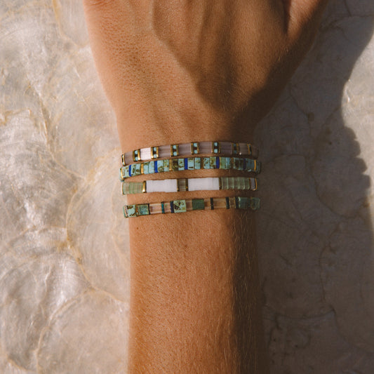 A close-up of a person's hand and wrist wearing multiple bracelets and rings on a light stone surface. The bracelets are made of green, blue, and white mosaic tiles, and the hand is decorated with gold rings, including a starfish-shaped ring.