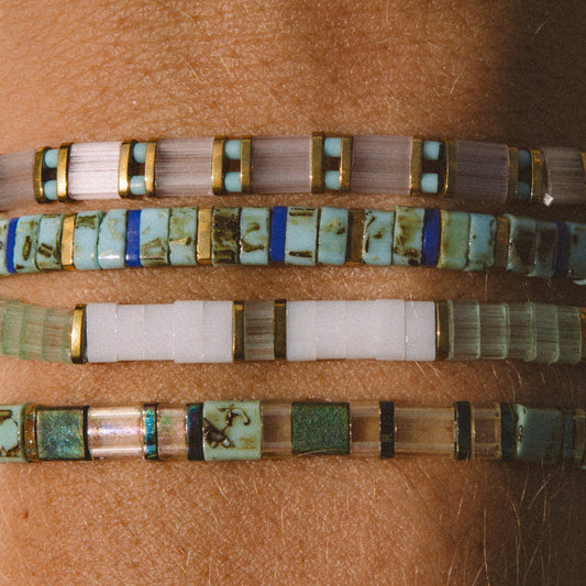 Four beaded bracelets worn stacked on a person's wrist, featuring turquoise, white, green, and brown inlay beads with gold-toned metal borders.