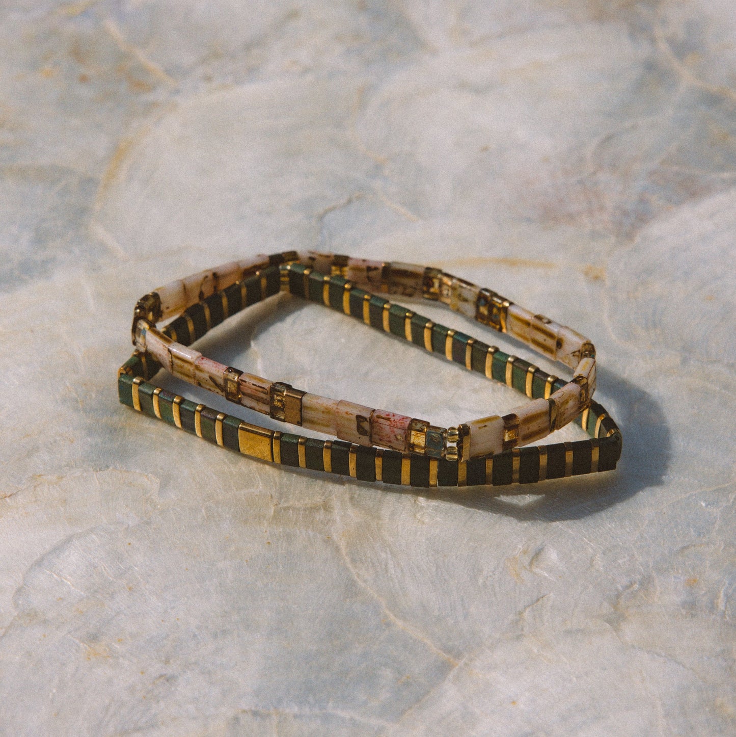A beaded stretch bracelet with a pattern of black, gold, and beige, displayed on a textured light gray stone surface.