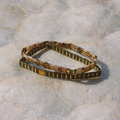 A beaded stretch bracelet with a pattern of black, gold, and beige, displayed on a textured light gray stone surface.