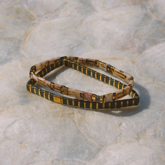 A beaded stretch bracelet with a pattern of black, gold, and beige, displayed on a textured light gray stone surface.
