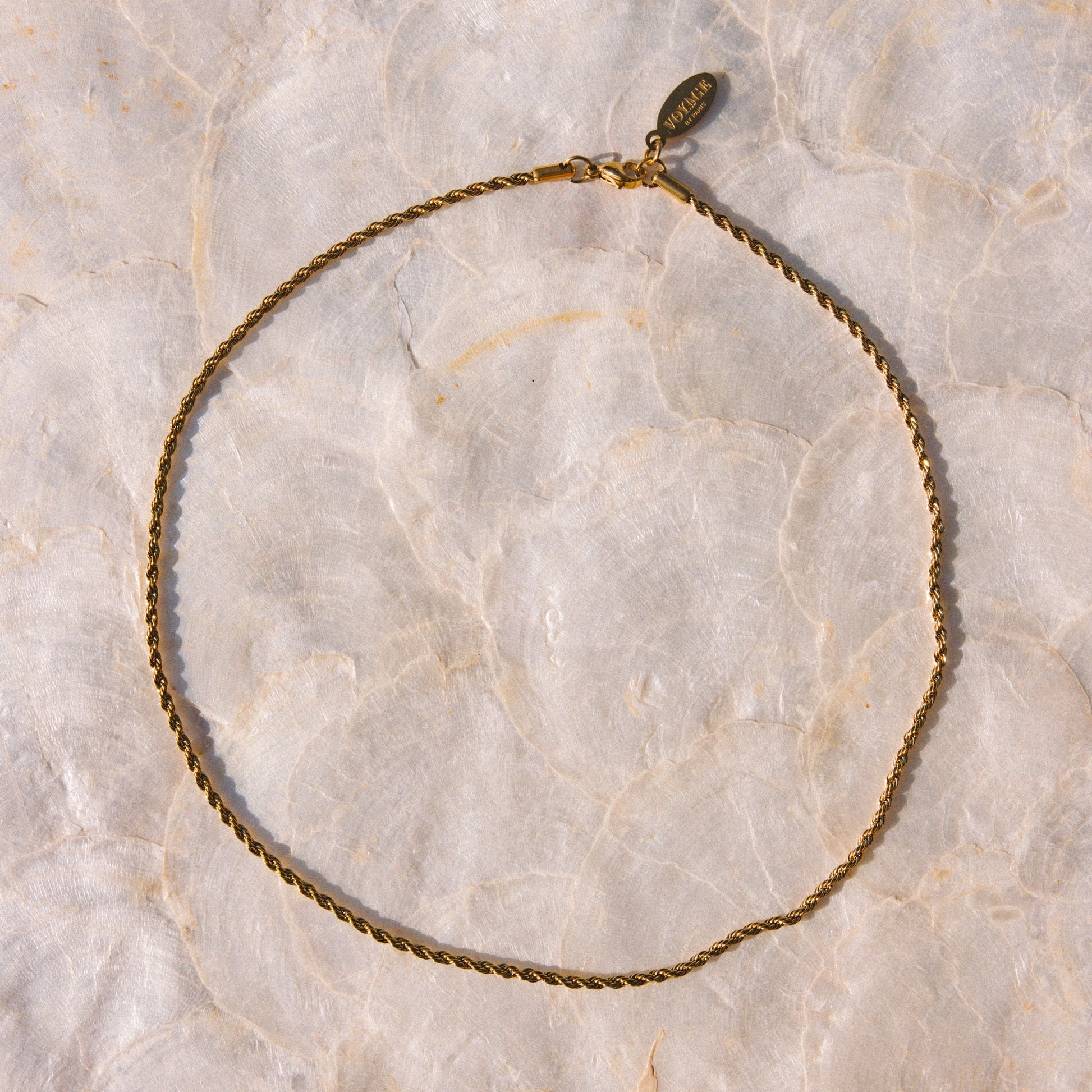 A delicate gold chain necklace with a small oval charm, laid flat on a textured beige marble surface.