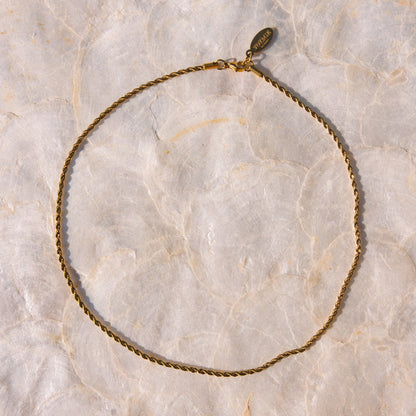A delicate gold chain necklace with a small oval charm, laid flat on a textured beige marble surface.