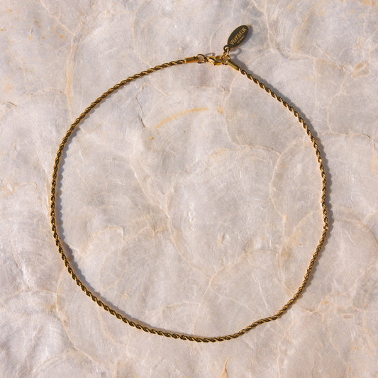 A delicate gold chain necklace with a small oval charm, laid flat on a textured beige marble surface.