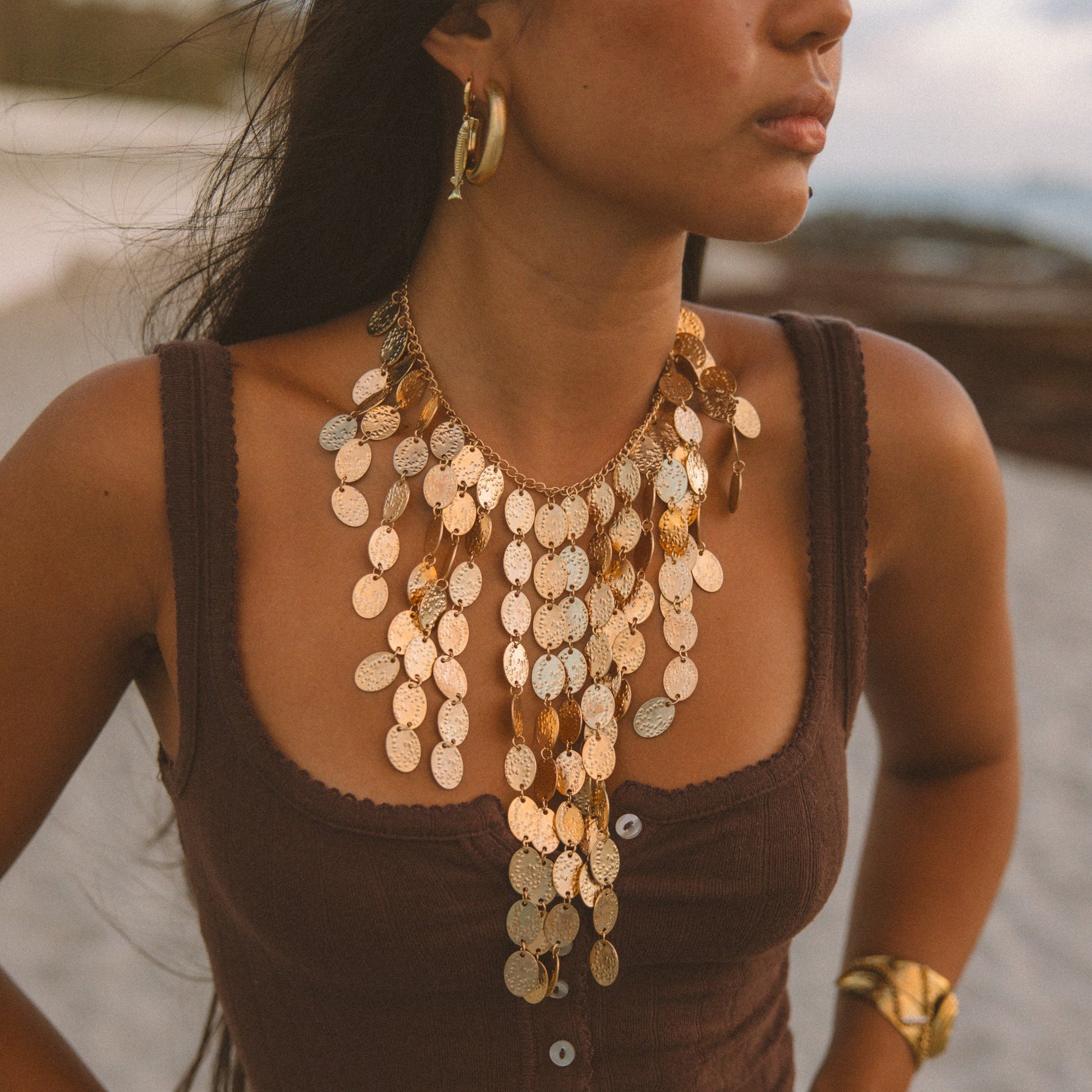 A woman wearing a layered bohemian gold coin necklace with disc-shaped charms, a matching gold hoop earring, and a gold bracelet, posing on a beach in a brown ribbed tank top.
