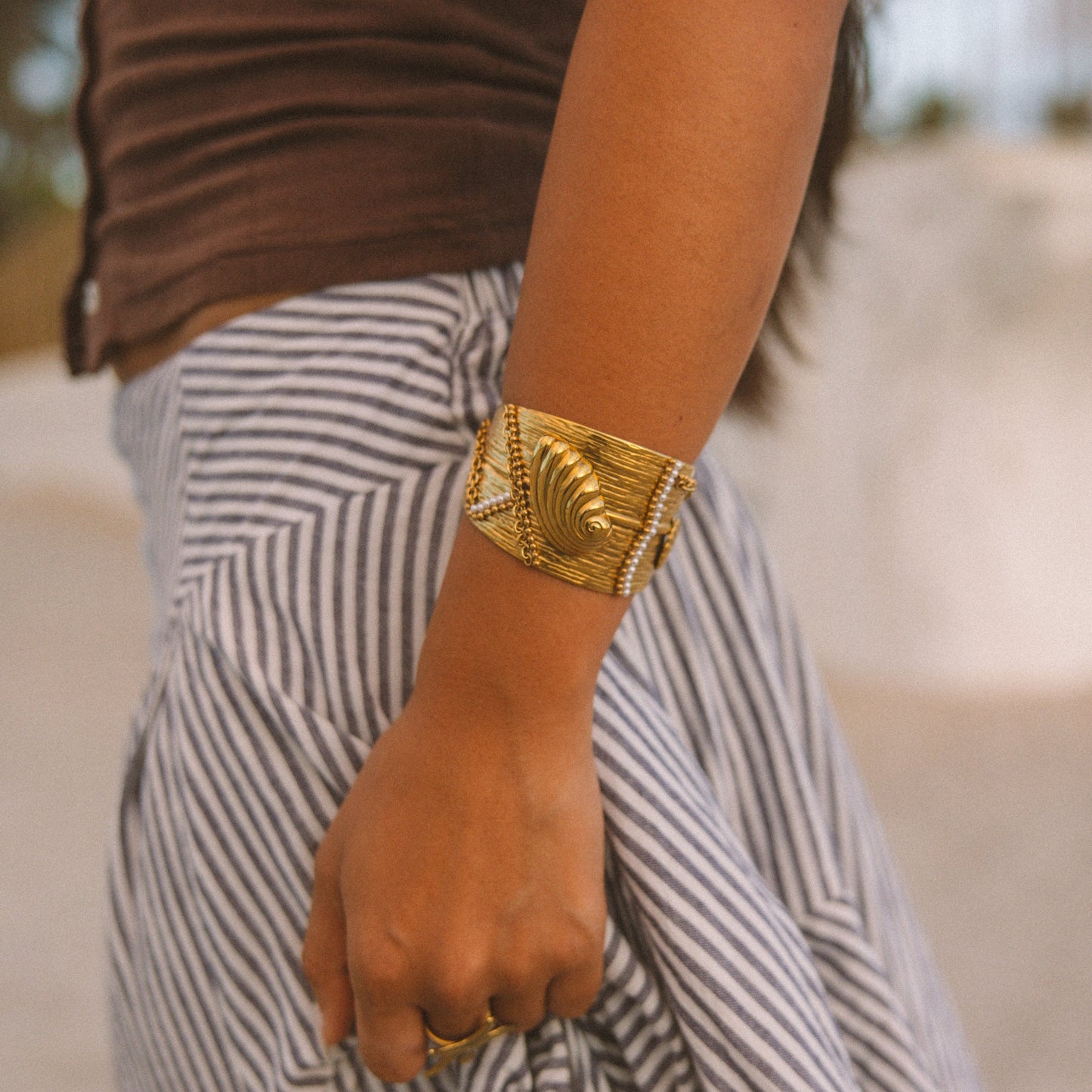 A close-up lifestyle shot of a person wearing a wide, chunky gold cuff bracelet on their wrist, paired with a brown top and black-and-white striped pants.
