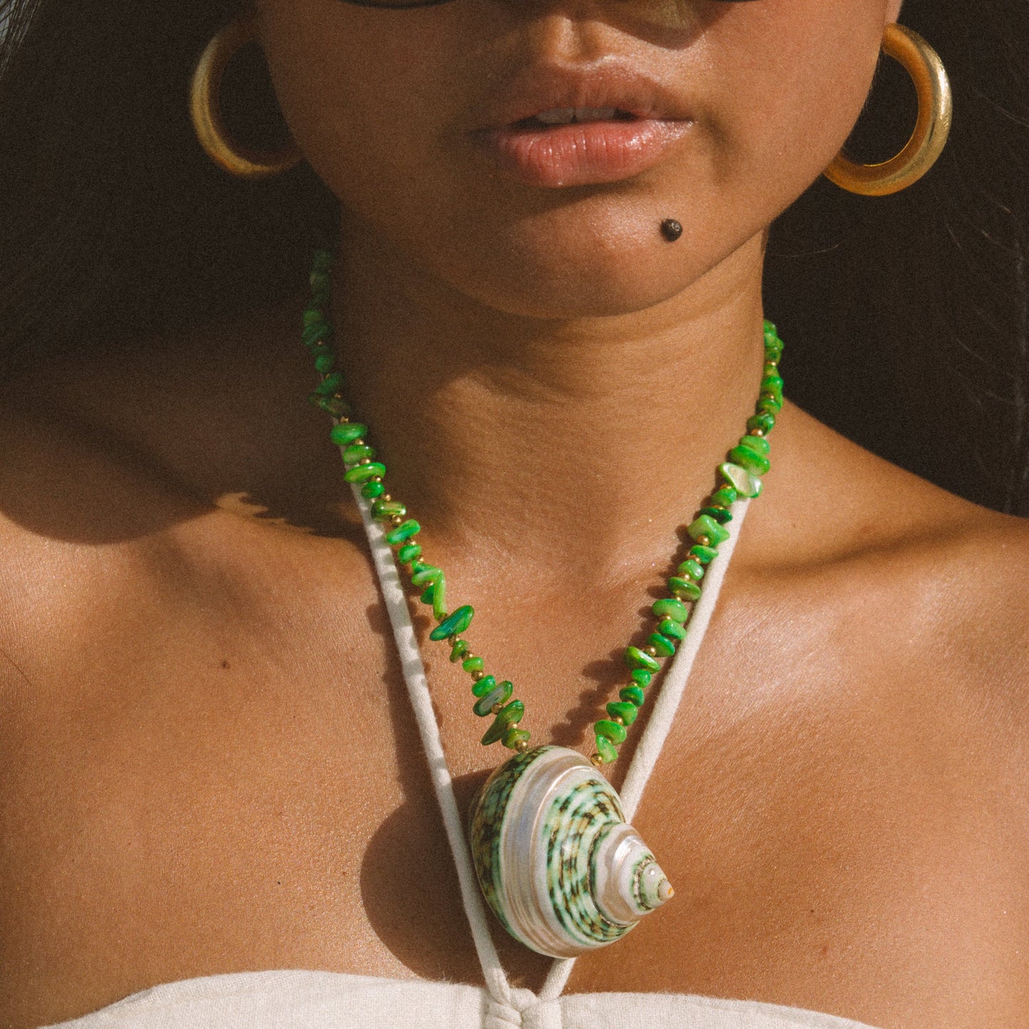 A close-up lifestyle shot of a model wearing a bohemian-style necklace with a large green and white seashell pendant, green chip beads, and white cord straps, styled with gold hoop earrings and sunglasses.