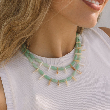 A close-up lifestyle shot of a woman wearing a double-strand statement necklace made of mint green beads and white pointed teeth, paired with a white ribbed tank top.