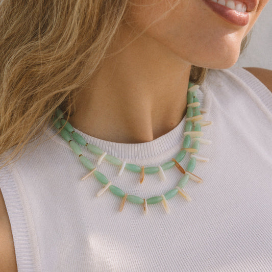 A close-up lifestyle shot of a woman wearing a double-strand statement necklace made of mint green beads and white pointed teeth, paired with a white ribbed tank top.