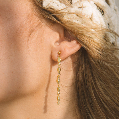 A close-up of a woman's ear wearing a delicate gold dangle earring with small faceted beads, against warm sunlight and a white knit headband.