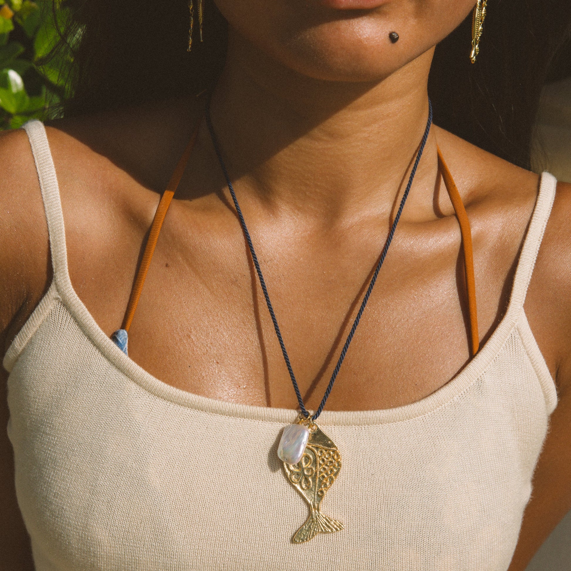 A close-up lifestyle shot of a woman wearing a gold fish pendant necklace on a black cord over a cream-colored ribbed tank top.