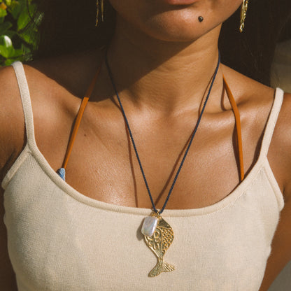 A close-up lifestyle shot of a woman wearing a gold fish pendant necklace on a black cord over a cream-colored ribbed tank top.