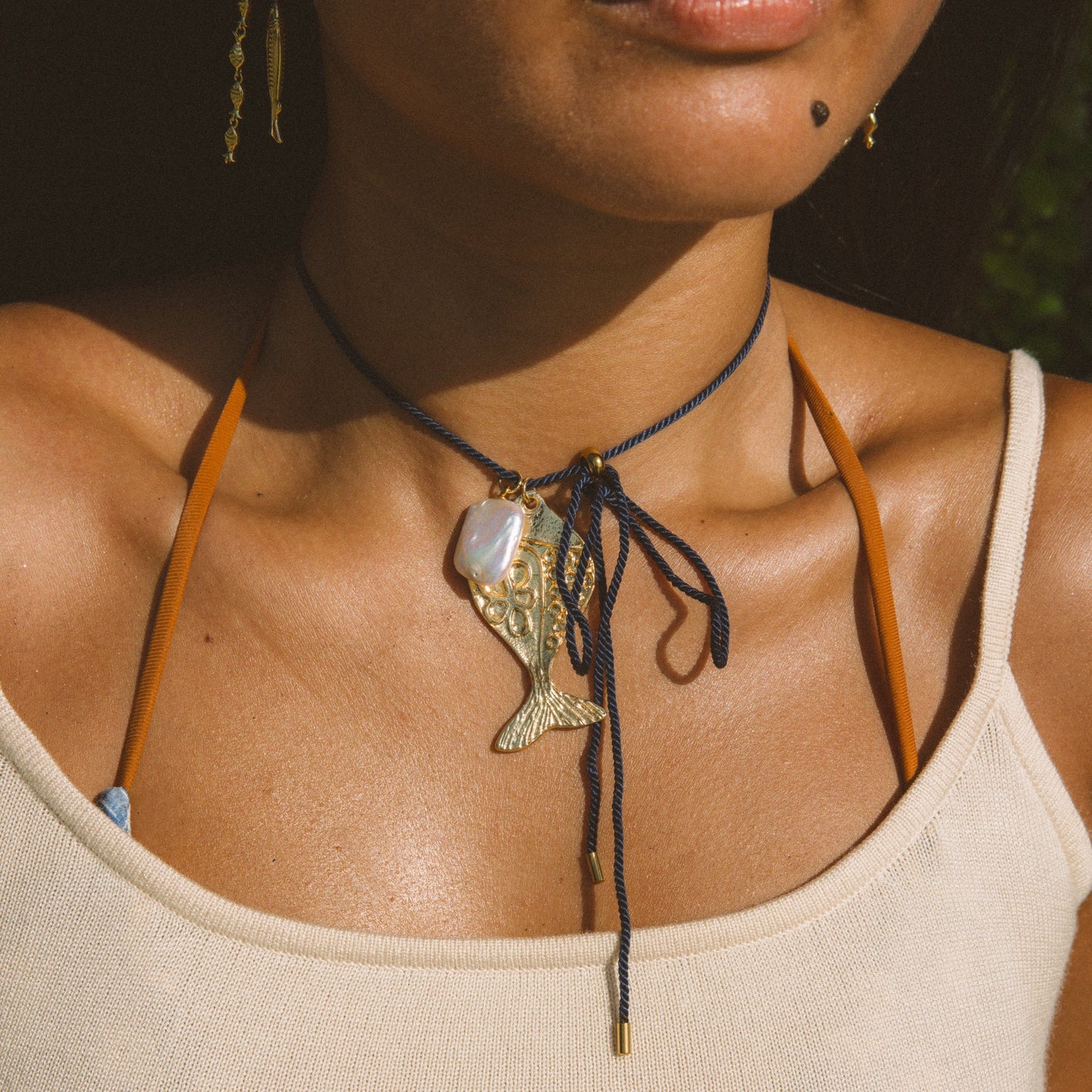 A close-up lifestyle shot of a model wearing a dark navy blue cord choker necklace with a gold-toned fish pendant and a white shell accent, styled with a cream ribbed tank top.
