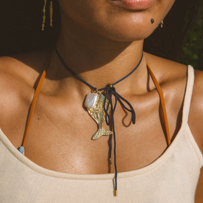 A close-up lifestyle shot of a model wearing a dark navy blue cord choker necklace with a gold-toned fish pendant and a white shell accent, styled with a cream ribbed tank top.